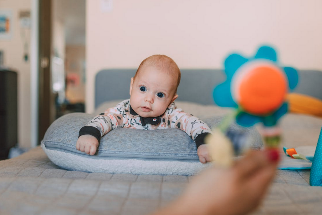 Baby laying on a pillow looking at a person behind the camera, an arm with a toy is in front of the camera